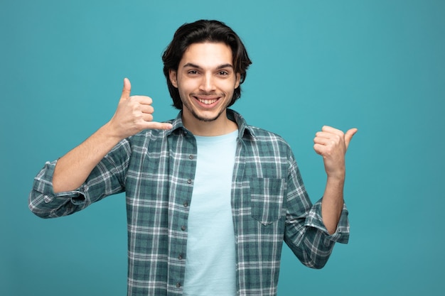 un joven apuesto sonriente mirando la cámara mostrando un gesto de llamada apuntando a un lado aislado de fondo azul