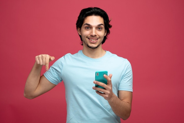 un joven apuesto sonriente sosteniendo un teléfono móvil mirando a la cámara apuntando hacia abajo aislado en un fondo rojo