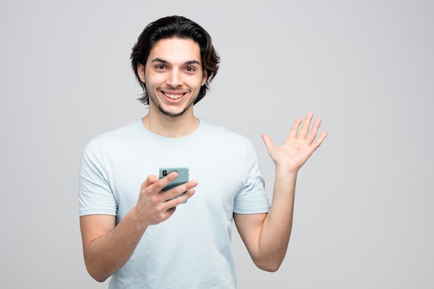 un joven apuesto sonriente sosteniendo un teléfono móvil mirando una cámara mostrando una mano vacía aislada de fondo blanco