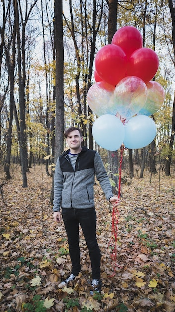 Un joven apuesto sostiene los globos de colores en un paseo por el bosque de otoño