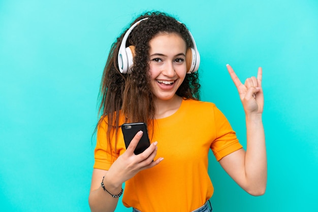 Joven árabe aislada de fondo azul escuchando música con un móvil haciendo gestos de rock