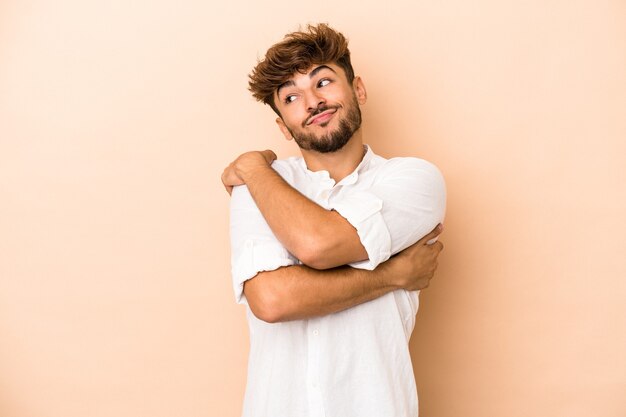 Joven árabe aislado sobre fondo beige abrazos, sonriendo despreocupado y feliz.