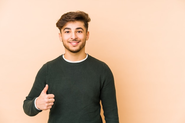 Joven árabe aislado sobre un fondo beige sonriendo y levantando el pulgar