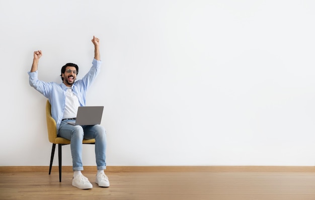 Un joven árabe alegre con ropa informal y gafas se sienta en un sillón con un portátil y hace un gesto de