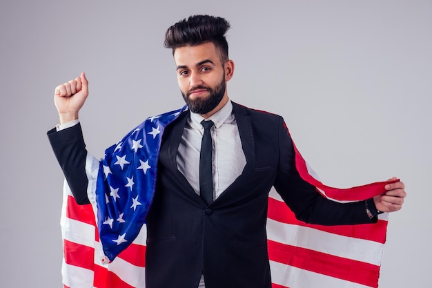 Joven árabe apuesto hombre de cabello negro con elegante traje de negocios que sostiene la bandera estadounidense en el estudio aislado fondo blanco Concepto del Día de la Independencia