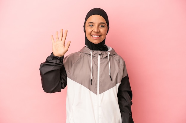 Joven árabe con burka de deporte aislado sobre fondo rosa sonriendo alegre mostrando el número cinco con los dedos.