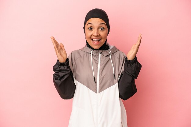 Joven árabe con burka deportiva aislada de fondo rosa recibiendo una agradable sorpresa emocionada y levantando las manos