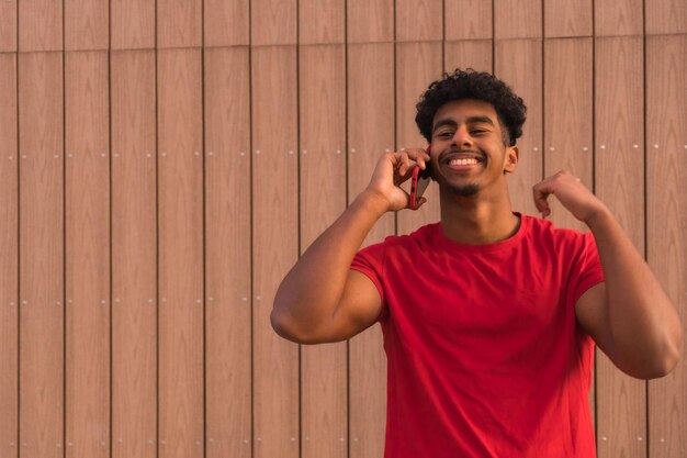 Joven árabe con camiseta roja en un fondo de madera sonriendo hablando por teléfono