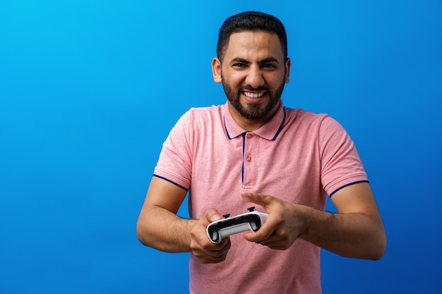 Joven árabe en camiseta rosa jugando videojuegos contra el fondo azul.