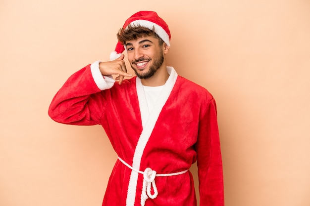 Joven árabe disfrazado de santa claus aislado sobre fondo beige mostrando un gesto de llamada de teléfono móvil con los dedos.