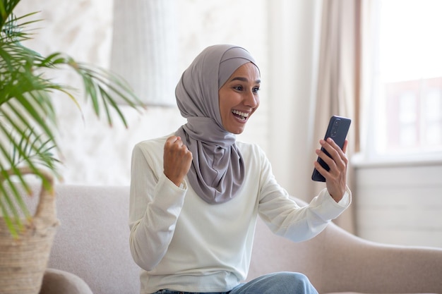 Una joven árabe feliz con hiyab sentada en un sofá en casa y usando un teléfono móvil mira la pantalla