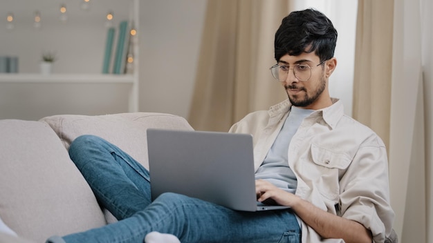 Joven árabe freelance hombre de negocios hombre barbudo estudiante con gafas yace en el sofá en casa con computadora portátil trabajando estudiando leyendo en línea pensando en la conexión problema frotando el mentón reflexionando solución