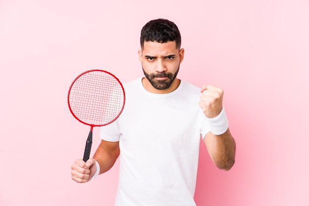 Joven árabe jugando bádminton aislado mostrando puño, expresión facial agresiva.