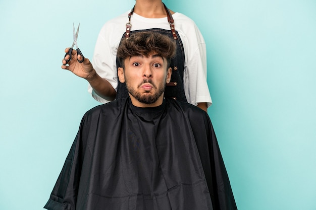 Joven árabe listo para cortarse el pelo aislado sobre fondo azul se encoge de hombros y abre los ojos confundidos.