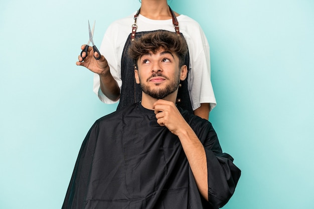 Joven árabe listo para cortarse el pelo aislado sobre fondo azul mirando hacia los lados con expresión dudosa y escéptica.