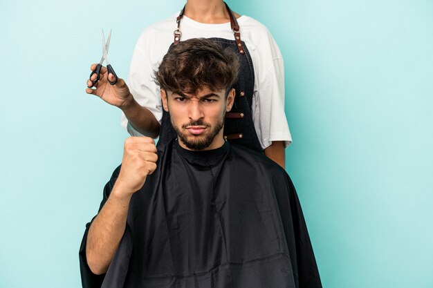 Foto joven árabe listo para cortarse el pelo aislado sobre fondo azul mostrando el puño a la cámara, expresión facial agresiva.