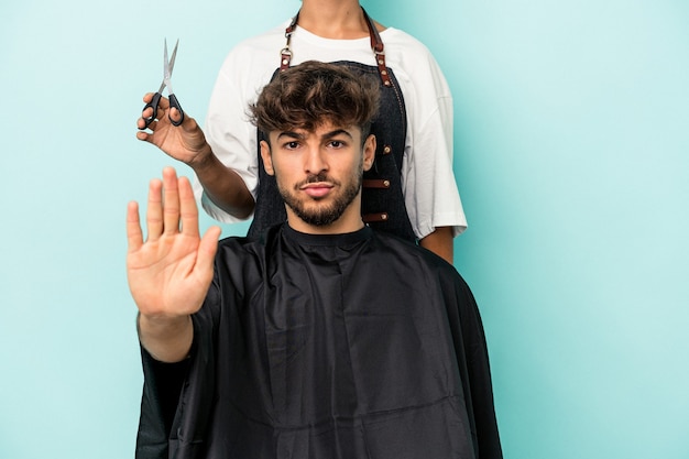 Joven árabe listo para cortarse el pelo aislado sobre fondo azul de pie con la mano extendida mostrando la señal de stop, impidiéndote.