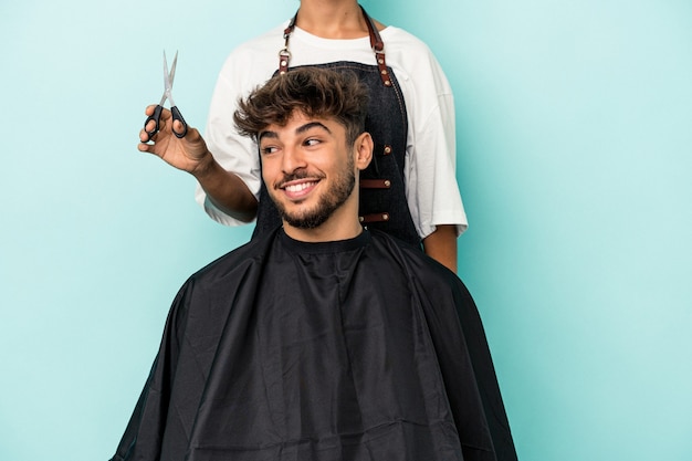 Joven árabe listo para cortarse el pelo aislado sobre fondo azul se ve a un lado sonriente, alegre y agradable.