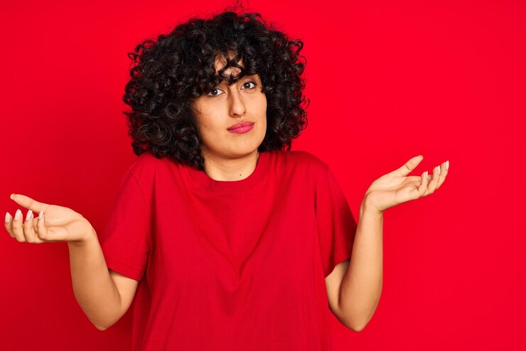 Foto joven árabe con el pelo rizado y una camiseta informal sobre un fondo rojo aislado expresión despistada y confusa con los brazos y las manos levantados concepto de duda