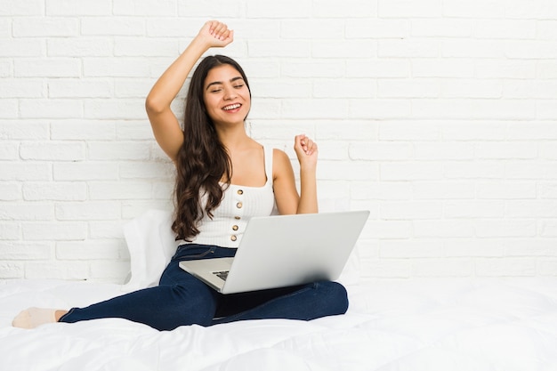 Joven árabe que trabaja con su computadora portátil en la cama celebrando un día especial, salta y levanta los brazos con energía.