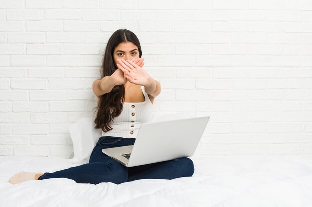 Joven árabe que trabaja con su computadora portátil en la cama haciendo un gesto de negación