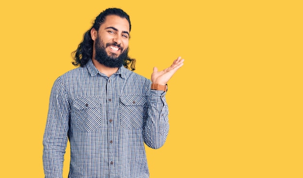 Joven árabe con ropa informal sonriendo alegre presentando y señalando con la palma de la mano mirando a la cámara