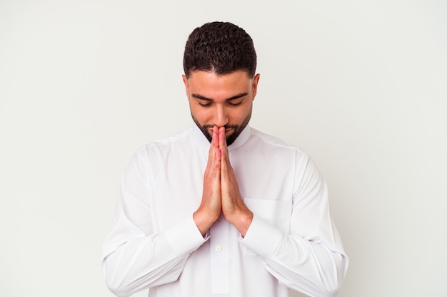 Foto joven árabe con ropa típica árabe aislado en la pared blanca tomados de la mano en oración cerca de la boca, se siente confiado.