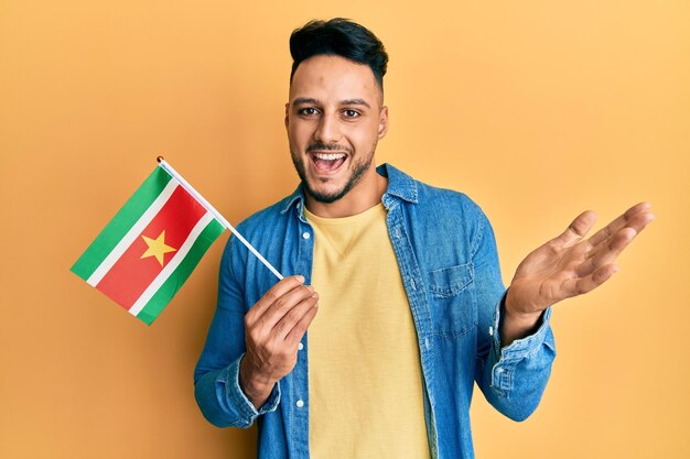 Joven árabe sosteniendo la bandera de surinam celebrando el logro con una sonrisa feliz y expresión ganadora con la mano levantada