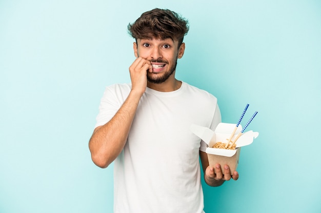 Joven árabe sosteniendo un fideos para llevar aislado sobre fondo azul mordiéndose las uñas, nervioso y muy ansioso.