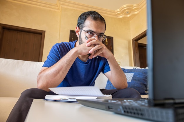 Joven árabe trabajando en casa con laptop y papeles en el escritorio