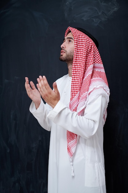 joven árabe vestido con ropa tradicional haciendo oración tradicional a Dios, mantiene las manos en gesto de oración frente a una pizarra negra que representa la moda islámica moderna y el concepto de ramadán kareem