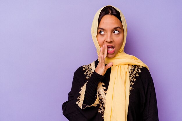 Joven árabe vistiendo un traje típico árabe aislado sobre fondo púrpura está diciendo una noticia secreta de frenado en caliente y mirando a un lado
