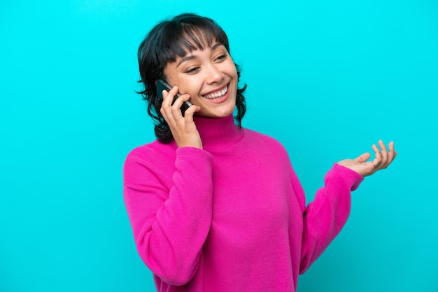 Joven argentina aislada de fondo azul manteniendo una conversación con alguien por teléfono móvil