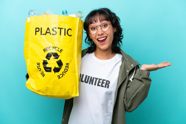 Joven argentina sosteniendo una bolsa llena de botellas de plástico para reciclar aisladas de fondo azul con una expresión facial conmocionada