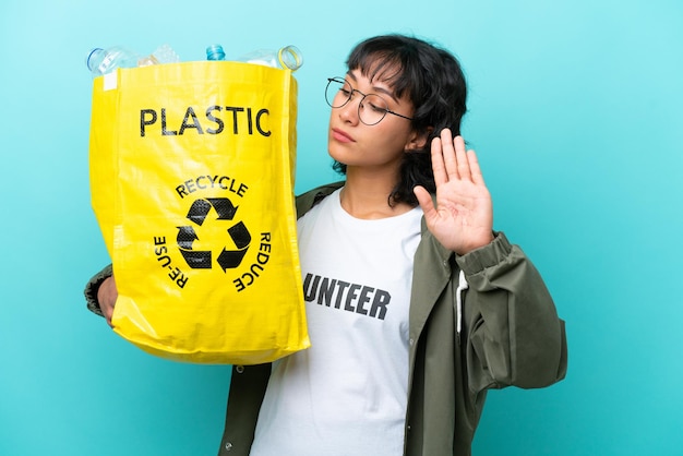 Joven argentina sosteniendo una bolsa llena de botellas de plástico para reciclar aisladas de fondo azul haciendo un gesto de parada y decepcionada