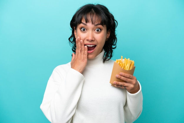 Joven argentina sosteniendo papas fritas aisladas de fondo azul con expresión facial sorprendida y sorprendida