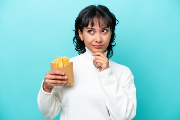 Joven argentina sosteniendo patatas fritas aisladas de fondo azul con dudas