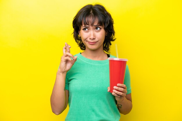 Joven argentina sosteniendo un refresco aislado de fondo amarillo con los dedos cruzados y deseando lo mejor