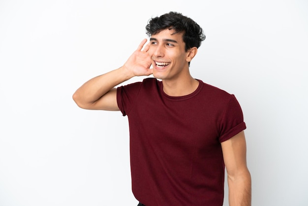 Joven argentino aislado de fondo blanco escuchando algo poniendo la mano en la oreja