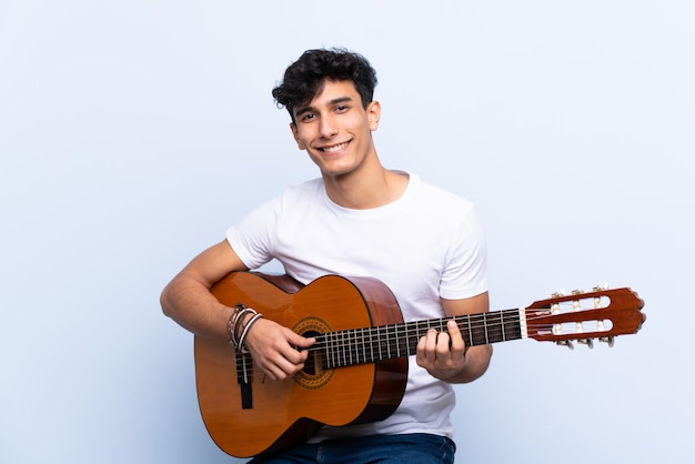 Joven argentino con guitarra sobre pared azul aislado mostrando signo bien con los dedos
