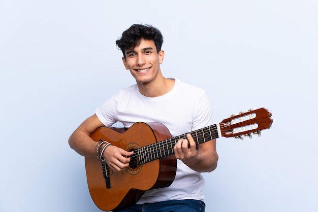 Joven argentino con guitarra sobre pared azul aislado sonriendo mucho