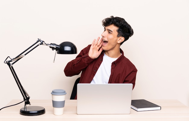 Joven argentino en una mesa con un portátil en su lugar de trabajo gritando con la boca abierta