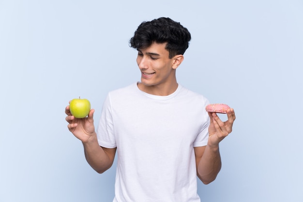 Joven argentino sobre pared azul aislada tomando una tableta de chocolate en una mano y una manzana en la otra