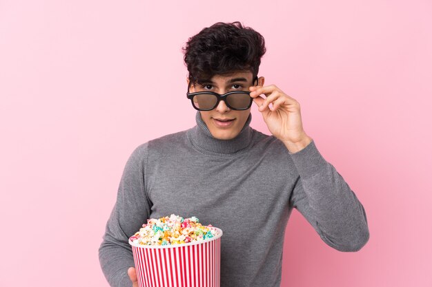 Joven argentino sobre pared blanca aislada sorprendido con gafas 3d y sosteniendo un gran cubo de palomitas de maíz