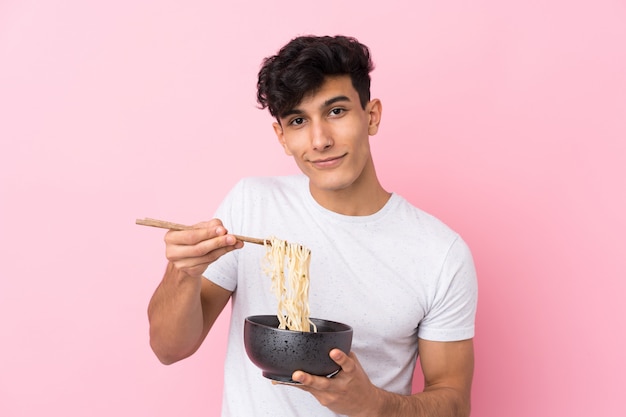 Joven argentino sobre pared blanca aislada sosteniendo un tazón de fideos con palillos y ofreciéndolo