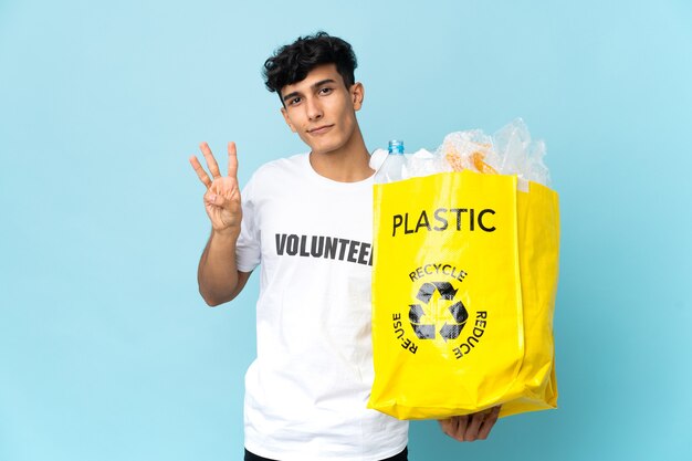 Joven argentino sosteniendo una bolsa llena de plástico feliz y contando tres con los dedos