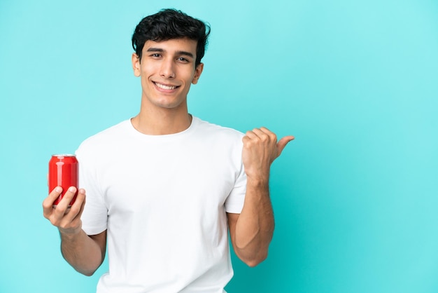 Joven argentino sosteniendo un refresco aislado de fondo azul apuntando hacia un lado para presentar un producto