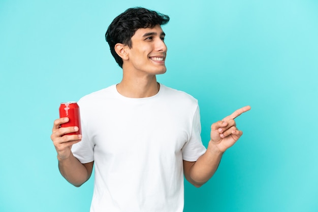 Joven argentino sosteniendo un refresco aislado de fondo azul señalando con el dedo al costado y presentando un producto