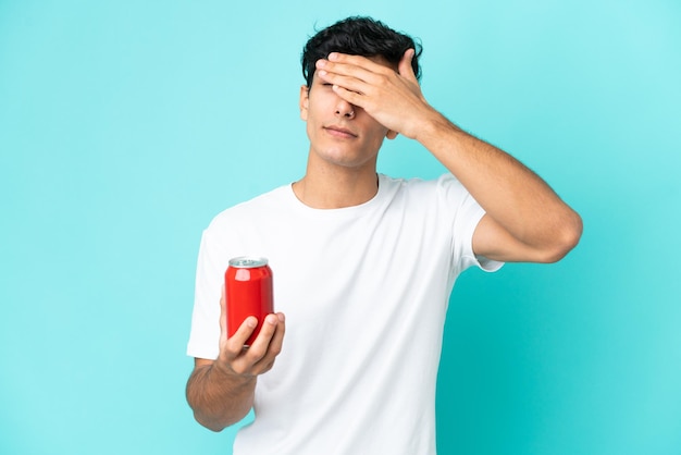 Joven argentino sosteniendo un refresco aislado sobre fondo azul cubriendo los ojos con las manos. No quiero ver algo