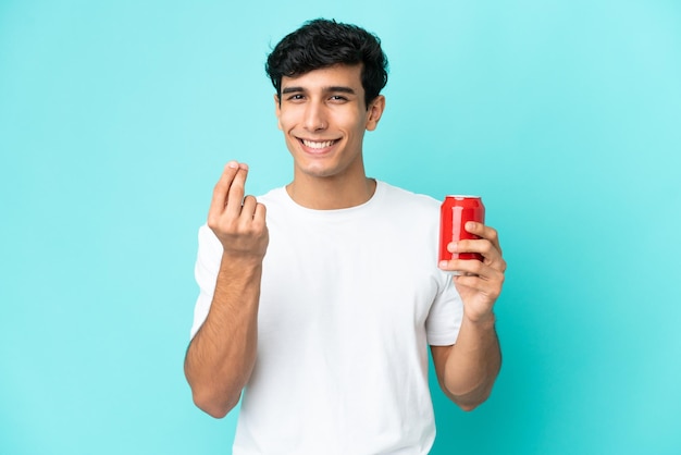 Joven argentino sosteniendo un refresco aislado sobre fondo azul haciendo gesto de dinero